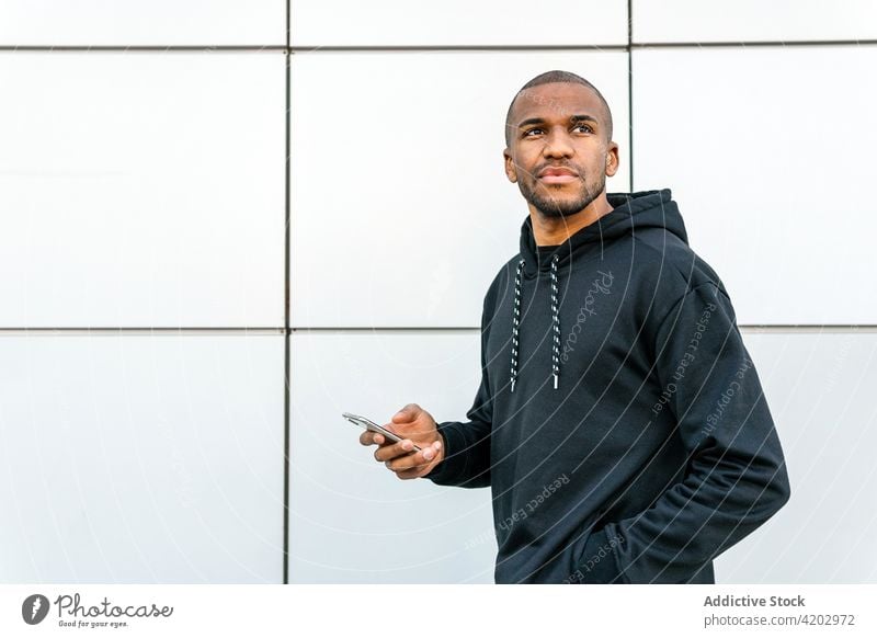 Maskuliner schwarzer Mann im Kapuzenpulli in der Nähe einer gekachelten Wand Hand-in-Tasche Stil maskulin Smartphone Browsen Fliesen u. Kacheln Stadt Großstadt