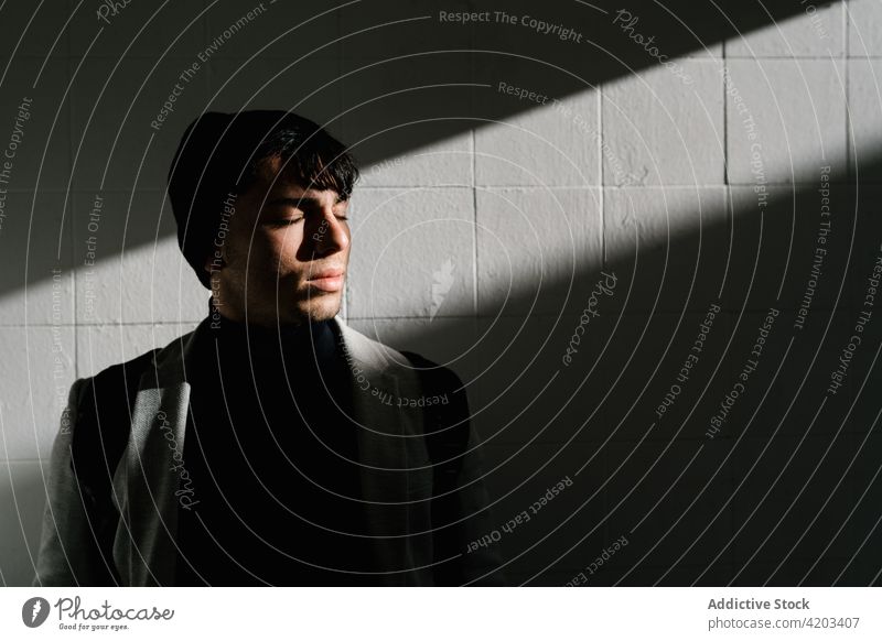 Stilvoller Mann steht in der Nähe der von Sonnenlicht beleuchteten Wand Gelassenheit Harmonie Schüler Schatten ruhig männlich Rucksack jung Großstadt urban