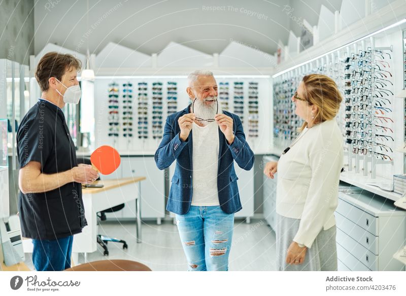 Lächelnder älterer Mann, der in einem Optikgeschäft eine Brille anprobiert anprobieren Wahl optisch Laden Optiker Kunde Spiegel Paar wählen Senior Zusammensein