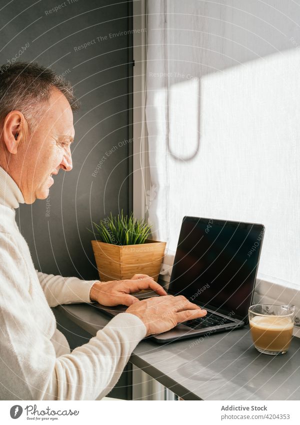 Fröhlicher reifer Mann sitzt auf einem Stuhl am Tresen mit Laptop in der Küche Morgen Lächeln heimwärts Frühstück Kaffee Abfertigungsschalter männlich Arbeit