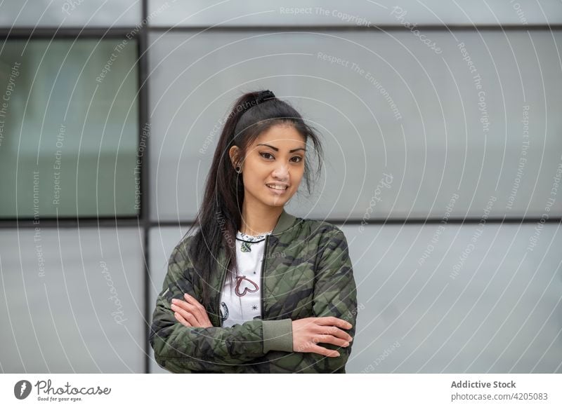 Selbstbewusste asiatische Frau auf einer Straße in der Stadt urban Stil Großstadt selbstbewusst die Arme verschränkt Outfit Gebäude Wand jung ethnisch trendy