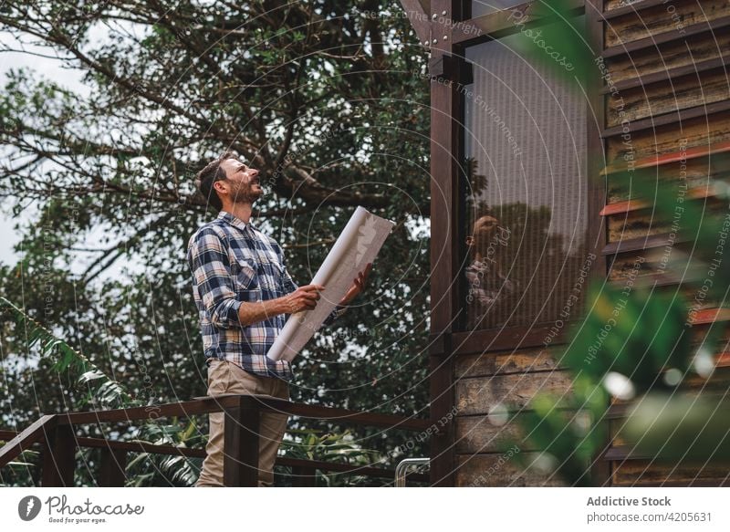 Männlicher Architekt mit Blaupause neben modernem Haus stehend Mann Bauplan Gebäude Projekt Tiefgang Papier Terrasse hölzern männlich Cottage Konstruktion Wald