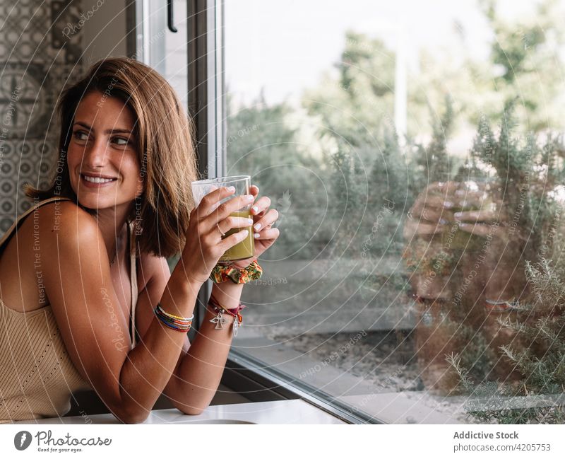 Ein kaukasisches Mädchen trinkt vor ihrem großen Küchenfenster einen grünen Gemüsesaft. Frau abgestimmt schütteln Layout Tischplatte Sellerie frisch Salat weich