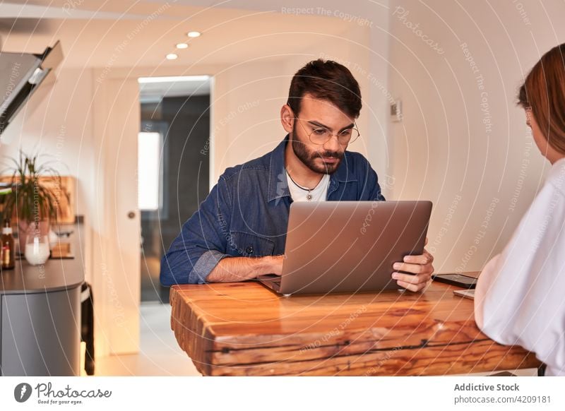 Ernster ethnischer Mann, der in der Küche in der Nähe seiner Freundin am Netbook arbeitet und ein Smartphone benutzt Paar freiberuflich Arbeit Laptop abgelegen