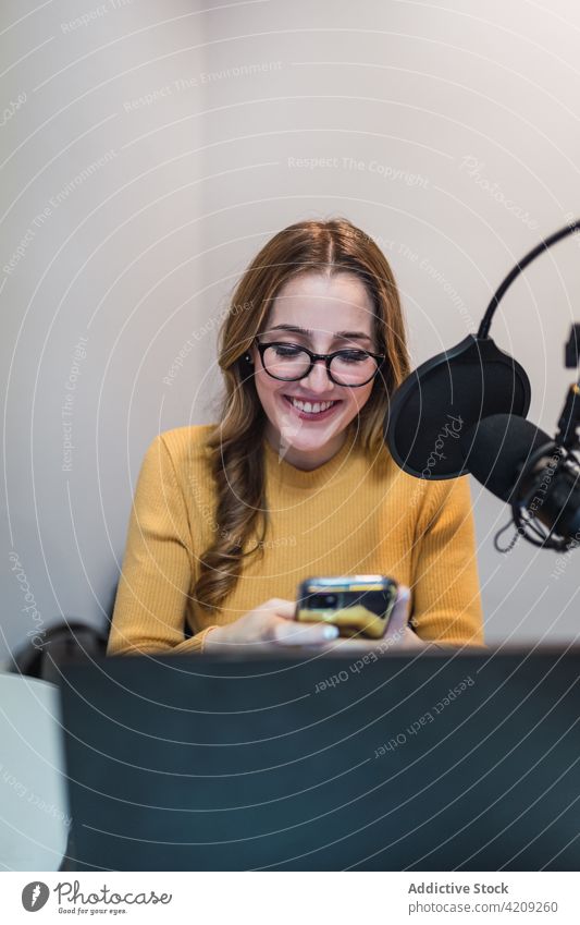 Zufriedene Frau, die im Radiosender ihr Smartphone durchsucht Textnachricht Arbeit Ausstrahlung Atelier Laptop Mikrofon Browsen Gerät Surfen modern SMS