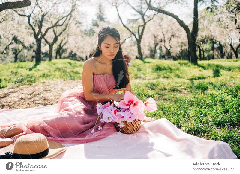 Ethnische Frau sitzend auf Plaid mit Blumen im Korb Blütezeit Rasen romantisch Obstgarten Frühling sinnlich friedlich träumen Garten Weide üppig (Wuchs) elegant