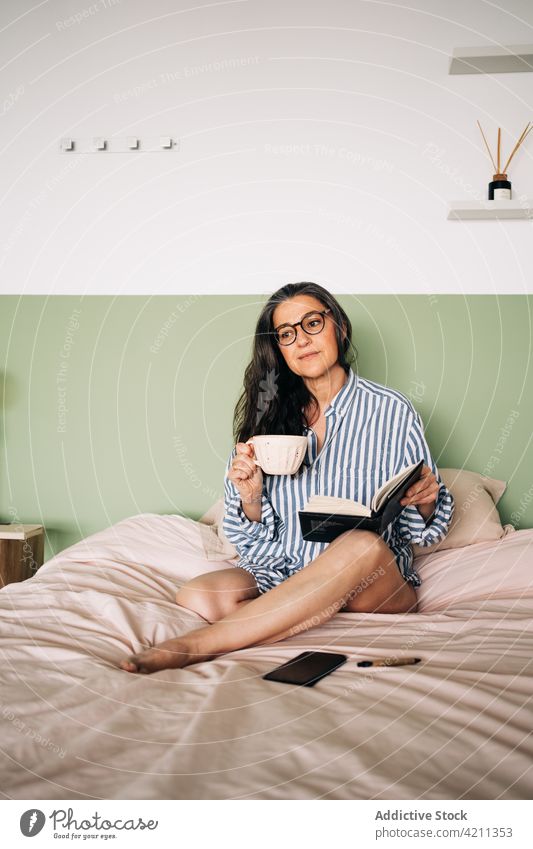 Frau mit Planer trinkt Kaffee auf dem Bett Tasse trinken Notebook Brille friedlich Freizeit Streifen Hemd heimwärts Schlafzimmer weich ruhig klug Komfort Dame