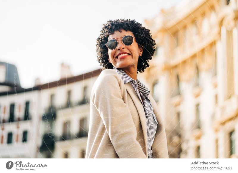 Fröhliche schwarze Frau mit trendiger Sonnenbrille lächelt auf der Straße heiter trendy Stadt Lächeln positiv Glück Mantel urban Mode Outfit Großstadt jung froh