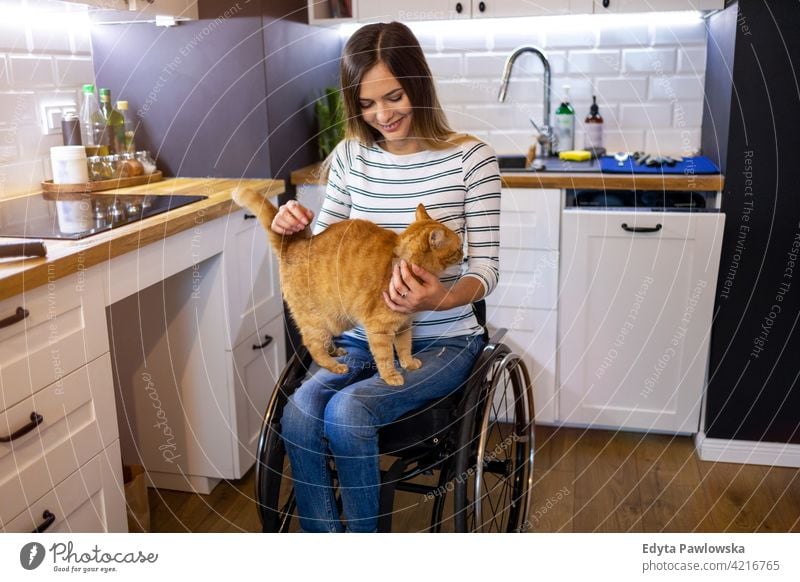 Behinderte junge Frau in der Küche mit Katze auf dem Schoß Rollstuhl häusliches Leben Behinderung deaktiviert Selbstvertrauen unabhängig im Innenbereich