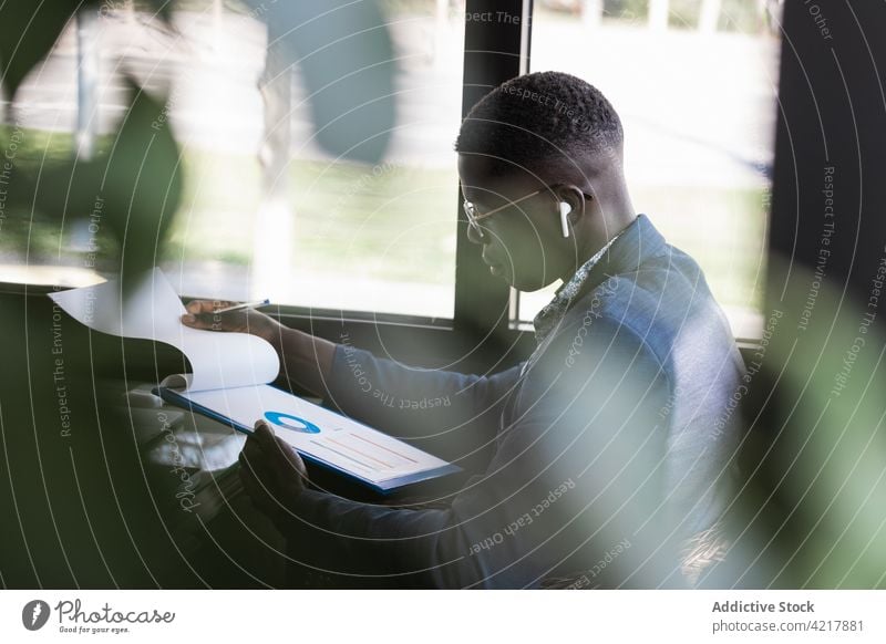 Seriöser schwarzer Geschäftsmann im Ohrhörer mit Diagramm auf Papier in einem Haus Arbeit Projekt Inbetriebnahme Ohrstöpsel ernst Zwischenablage Kantine