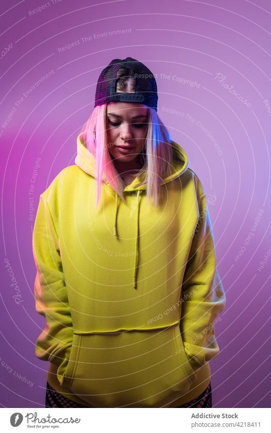 Coole Frau in stylischem Kapuzenpulli schaut im Studio weg Kokette Streetstyle neonfarbig Licht Atelier glühen Hut Verschlussdeckel Stil Outfit cool lebhaft