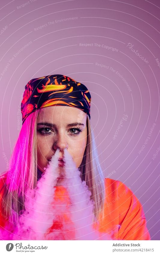 Frau raucht Vape im Studio mit Neonlicht Raps Rauch neonfarbig Atelier Licht e Zigarette cool Streetstyle Dunst Nikotin Habitus ungesund Mode ausatmen Stil