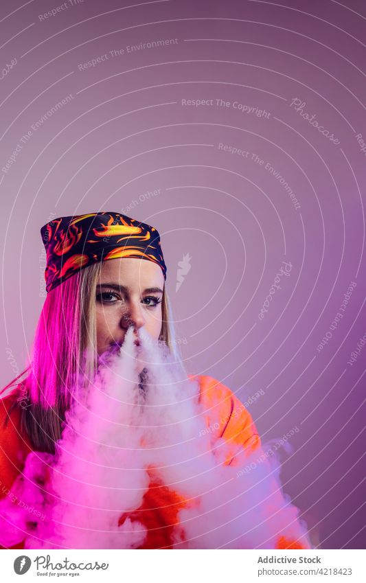 Frau raucht Vape im Studio mit Neonlicht Raps Rauch neonfarbig Atelier Licht e Zigarette cool Streetstyle Dunst Nikotin Habitus ungesund Mode ausatmen Stil