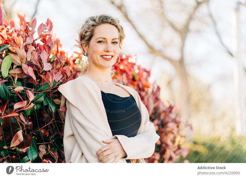 Stilvolle lächelnde Frau, die eine Gasse im Park entlanggeht Lächeln Spaziergang Kleid elegant Anmut Garten Baum Glück Mantel stehen charmant Natur trendy