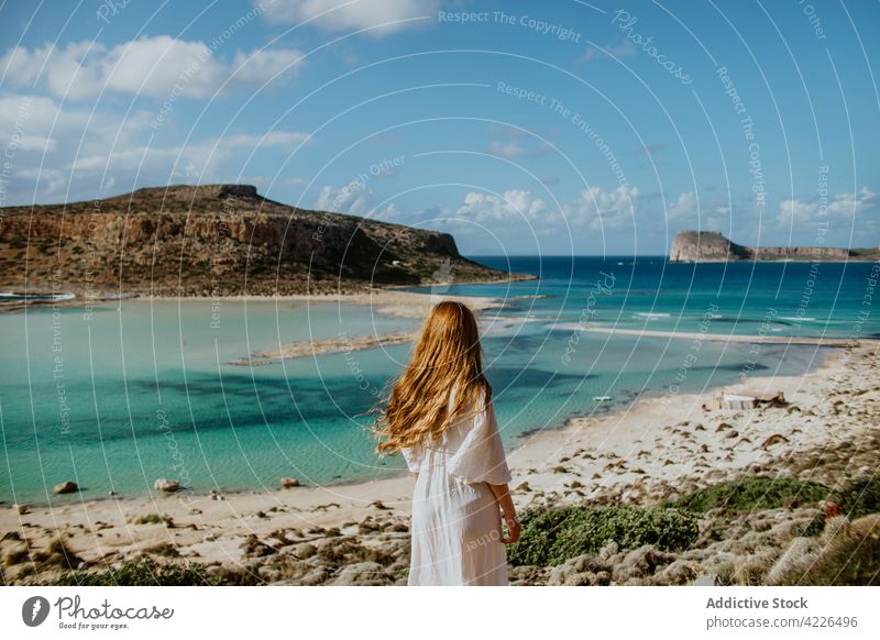 Unerkennbare Frau steht auf einem steinigen Aussichtspunkt vor azurblauem Meer Balos Strand Haare berühren Bergkuppe Strandbekleidung Resort Paradies malerisch