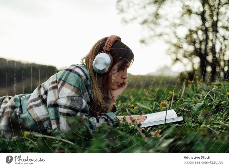 Frau mit Kopfhörern liest ein Buch im Gras lesen Literatur Wissen freie Zeit zuhören benutzend Apparatur Gerät Lehrbuch Bildung Musik Melodie tausendjährig