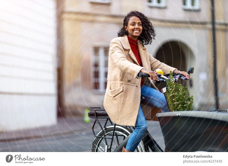 Junge Frau auf einem Lastenfahrrad in der Stadt mit kleinen Weihnachtsbaum in einem Kofferraum Weihnachten Weihnachtsbeleuchtung Weihnachtsdekorationen