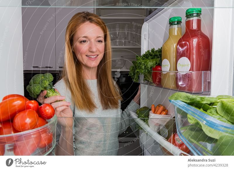 Offener Kühlschrank von innen, Glasregale mit buntem Gemüse, Flaschen mit Bio-Säften. Junge Frau mit glücklichem Lächeln nimmt Brokkoli. Gesunde Ernährung, veganes Konzept.