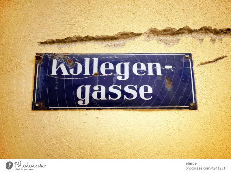 Beschädigtes, blau emailliertes Straßenschild " Kollegengasse " an einer ebenfalls beschädigten gelben Hauswand / wohnen Straßenname Schild Neid Wut kaputt