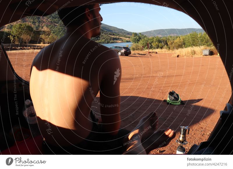 Warten mit Aussicht auf Landschaft und Kaffee - oder - junger Mann sitzt im schattigen Auto und schaut auf einen Parkplatz, während der Espresso kocht warten