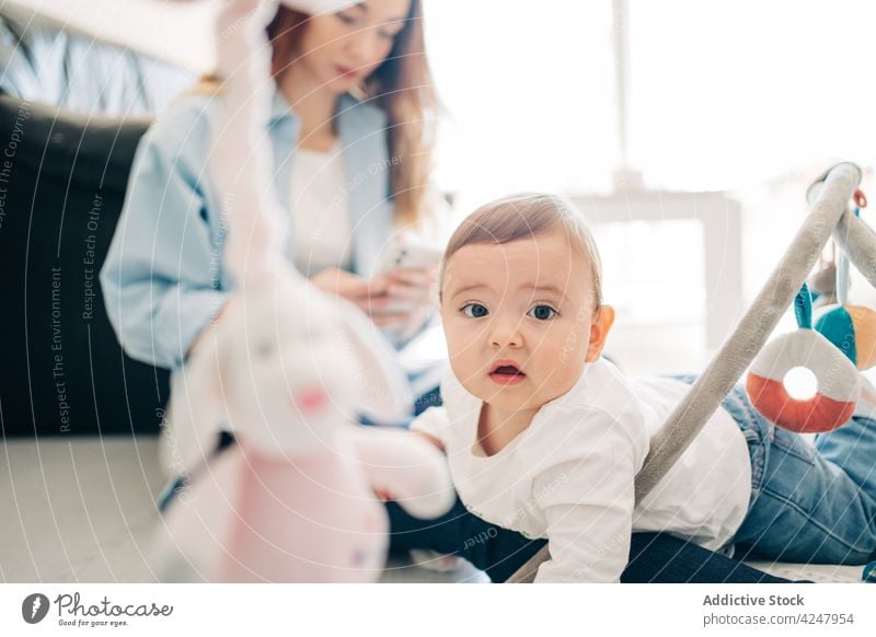 Lustiges Baby spielt auf dem Boden in der Nähe der Mutter, die auf ihrem Smartphone surft benutzend niedlich spielen Apparatur Säuglingsalter süß zu Hause