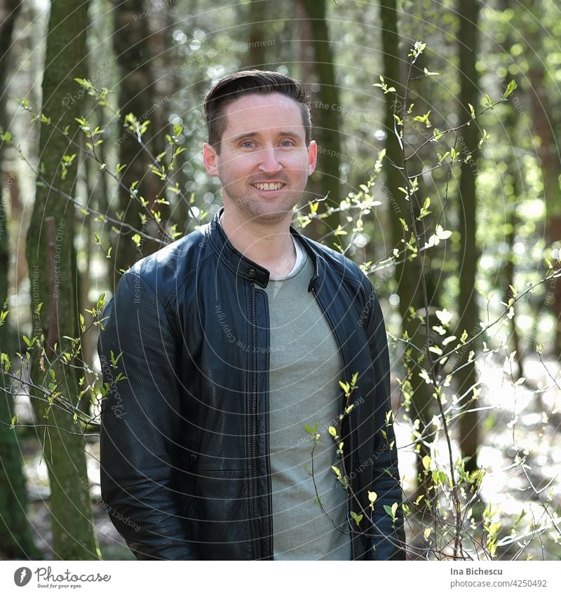 Ein junger Mann in Wald, neben einem dünnen Strauch mit kleine , hell grünen Blätter die von der Sonne beleuchtet werden. mann sportlich natur liebe blaue augen