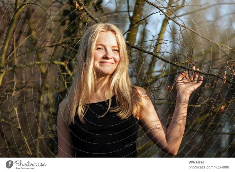 Ein lachendes blondes Mädchen spielt mit den Zweigen eines Baumes und freut sich über die  Sonne im Wald hübsch attraktiv fröhlich heiter lächeln froh glücklich