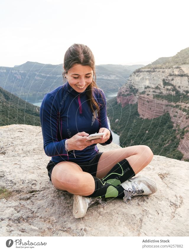 Fröhliche junge ethnische Frau lächelt und benutzt ihr Smartphone auf einer felsigen Klippe Nachricht Lächeln Schlucht Reisender Trekking Berge u. Gebirge