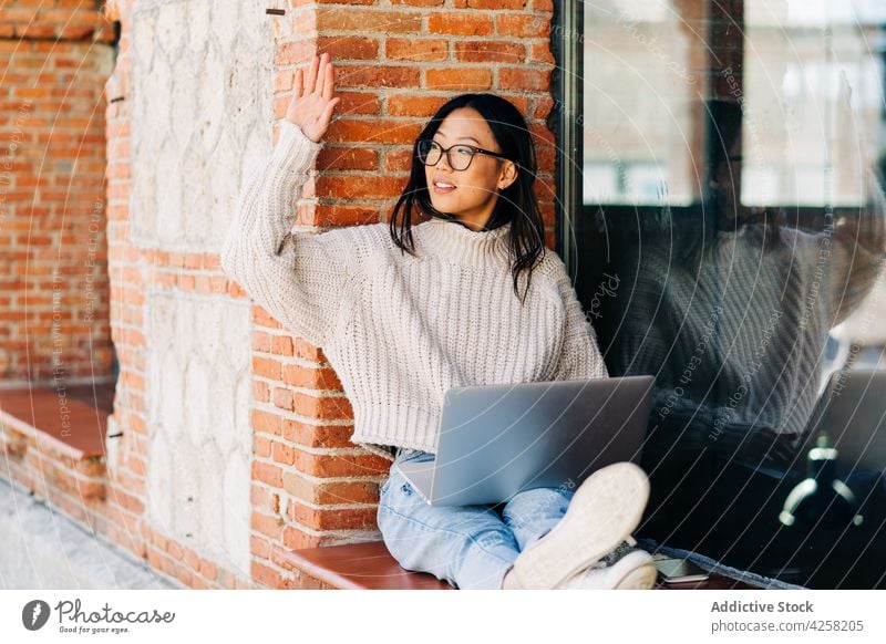 Trendige junge ethnische Dame grüßt Freundin bei der Arbeit am Laptop auf der Straße Frau Wellenhand Gruß abgelegen trendy Internet freiberuflich Sitzung