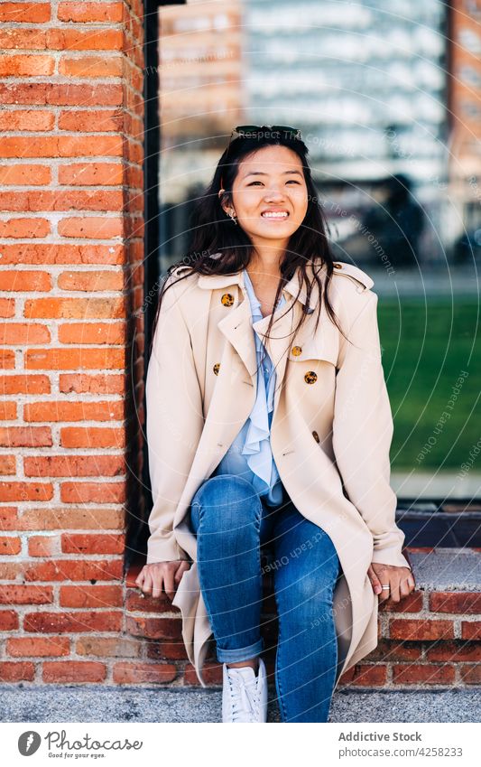 Trendy junge asiatische Frau sitzt in der Nähe von Backstein-Gebäude in der Stadt Straße trendy Stil selbstbewusst Persönlichkeit charismatisch positiv