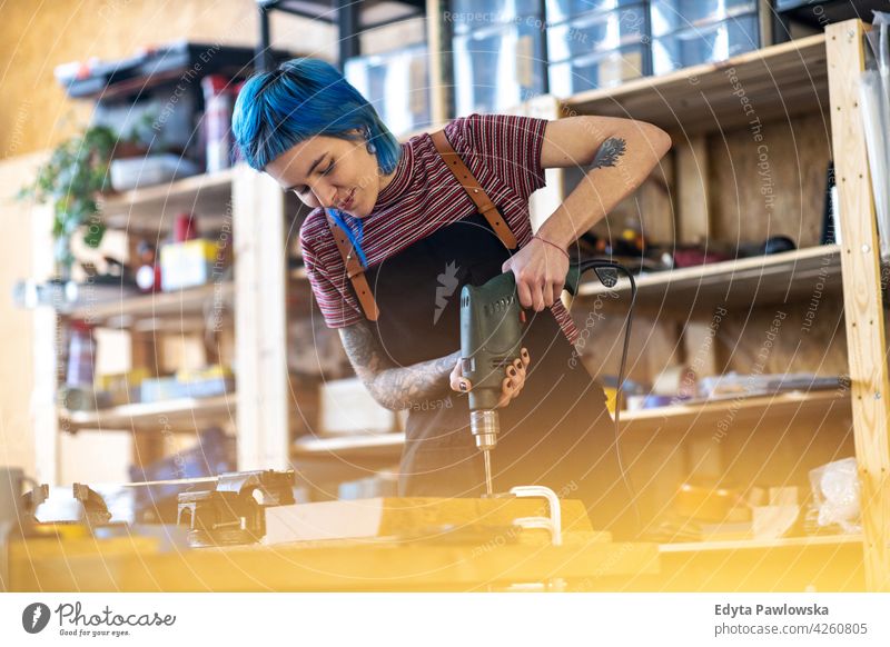 Junge Frau mit elektrischer Bohrmaschine in der Industriewerkstatt diy Hipster farbenfroh Behaarung Tattoos Besitzer Beruf Dienst Werkstatt Kleinunternehmen