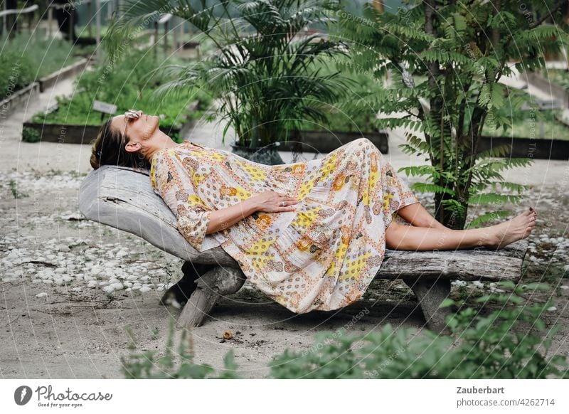 Frau im Kleid liegt entspannt auf einer Holzliege in einem Garten schön liegen entspannen Liege Urlaub chillen relaxen Sommer Reise Gemüsebeet Pflanzen grün