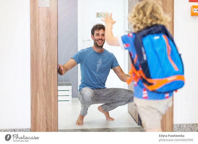 Kleiner Junge läuft auf seinen Vater zu, der zu Hause auf ihn wartet Bildung Schule Rucksack verlassend Morgen Eingangstür Saal echte Menschen anhänglich