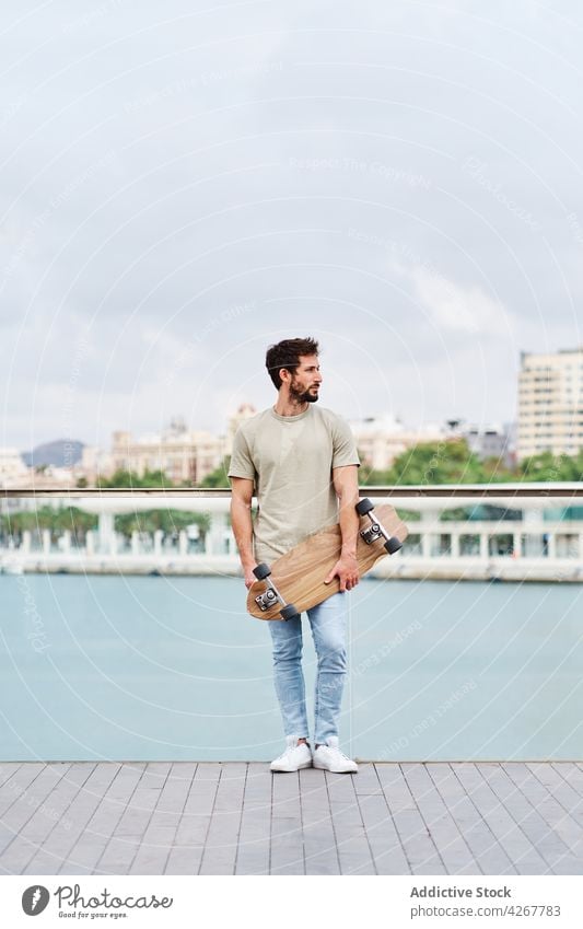 Bärtiger Mann mit Skateboard am Seeufer einer Stadt Skater Großstadt emotionslos lässig Hafengebiet Spazierweg Stauanlage jung Fluss Teich Sommer sonnig