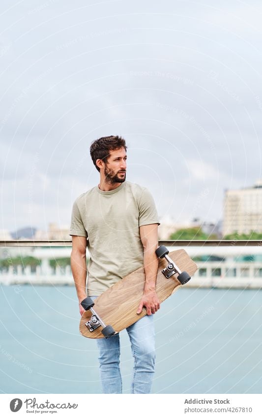 Bärtiger Mann mit Skateboard am Seeufer einer Stadt Skater Großstadt emotionslos lässig Hafengebiet Spazierweg Stauanlage jung Fluss Teich Sommer sonnig