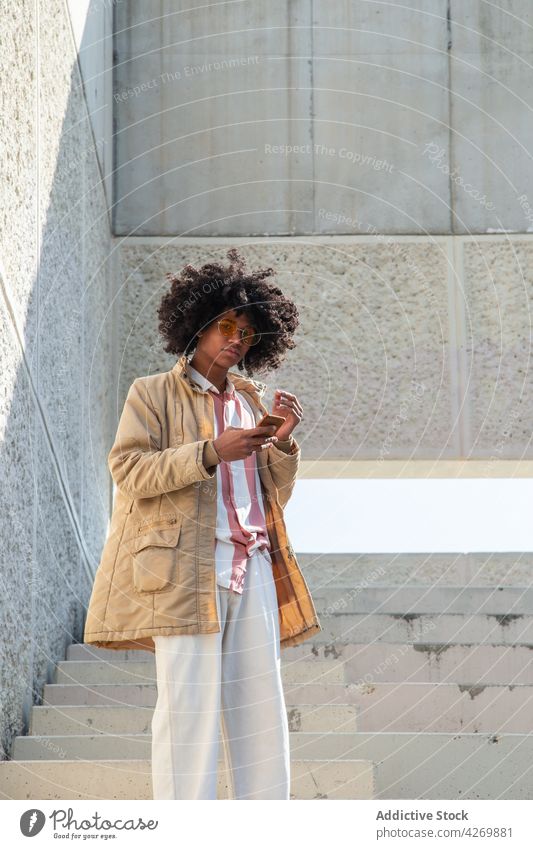 Schwarzer Mann in Retro-Kleidung plaudert auf einer Treppe mit seinem Smartphone Textnachrichten Hand-in-Tasche altehrwürdig Stil Afro-Look Internet benutzend