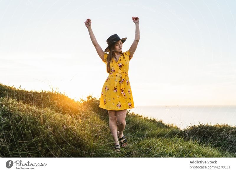 Junge Frau mit erhobenen Armen auf grasbewachsenem Rasen stehend Sonnenkleid Arme hochgezogen Landschaft Natur verträumt Grasland Wiese jung Feld Hut Vorschein