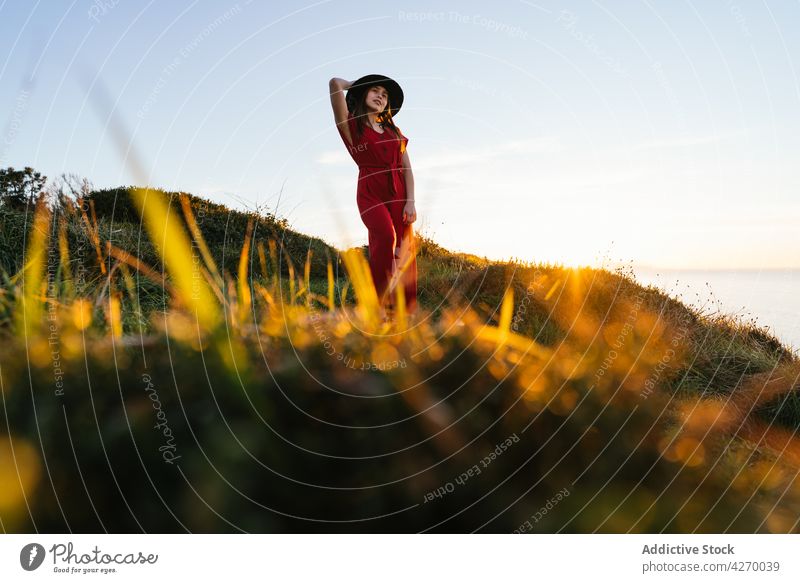 Junge Frau steht auf einem Rasenstück Sonnenkleid Landschaft Natur verträumt rot Grasland Wiese grasbewachsen jung Feld Hut Vorschein Gelassenheit Anmut feminin