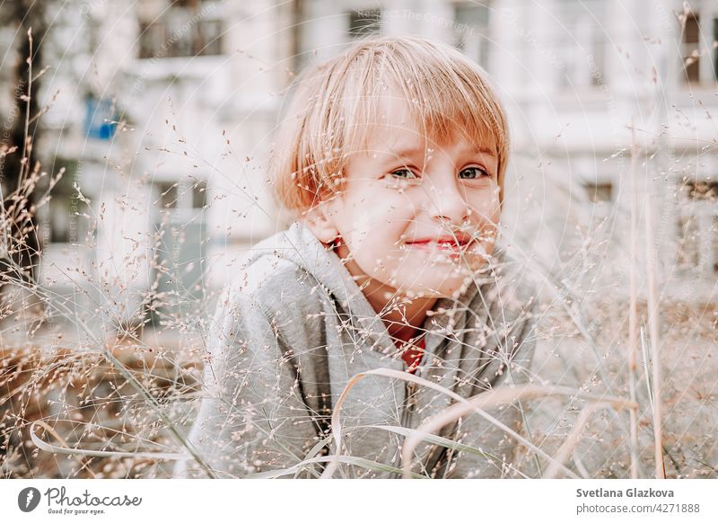 Porträt niedlich kühlen blonden kaukasischen Glücklich lächelnden kleinen Jungen in lässigen grauen Hoodie sitzen im hohen Gras vor dem Hintergrund der Natur.