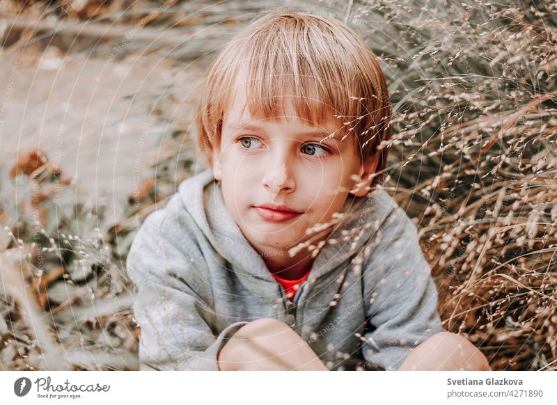 Porträt niedlich kühlen blonden kaukasischen Glücklich lächelnden kleinen Jungen in lässigen grauen Hoodie sitzen im hohen Gras vor dem Hintergrund der Natur.