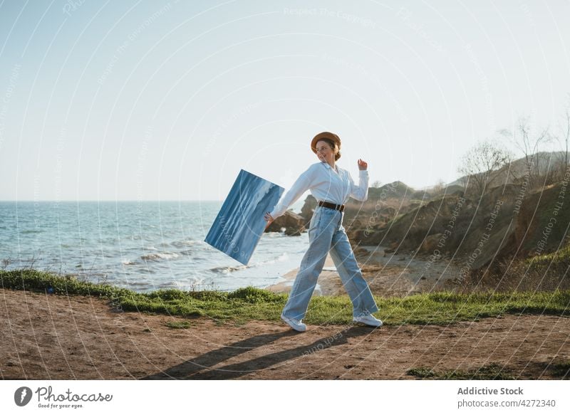 Lächelnde junge Frau, die mit einem Gemälde in der Hand am Meer spazieren geht Spaziergang Meeresufer Malerei Natur Inhalt Glück Anstreicher Strand Stil positiv