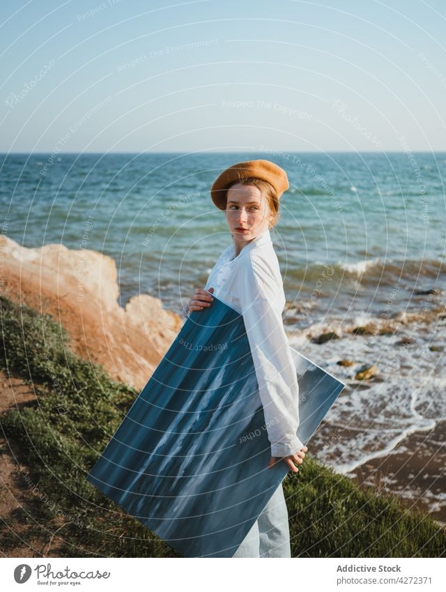 Junge Frau, die mit einem Gemälde in der Hand am Meer spazieren geht Spaziergang Meeresufer Malerei Natur Anstreicher Strand Stil MEER Küste jung trendy Hut