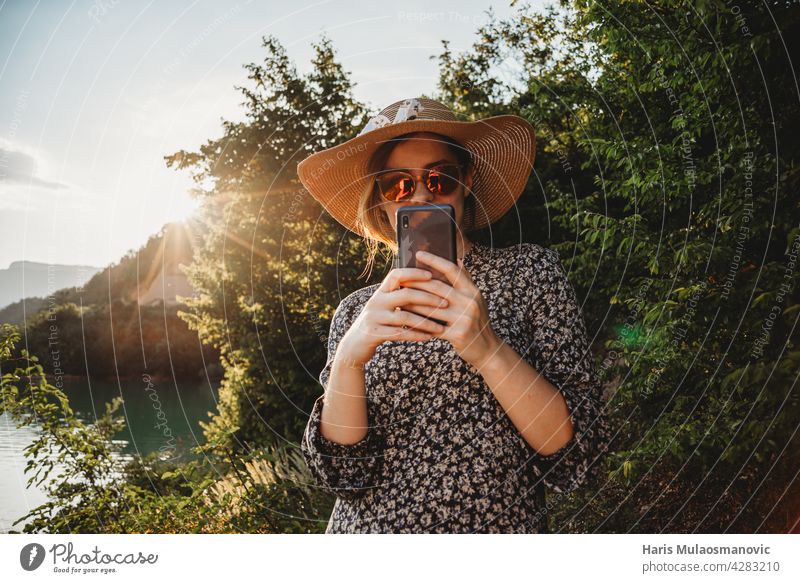 Frau mit Hut und Sonnenbrille mit Handy im Sonnenuntergang auf der Seeseite 5g Erwachsener App attraktiv Hintergrund schön Schönheit Kaukasier Mitteilung