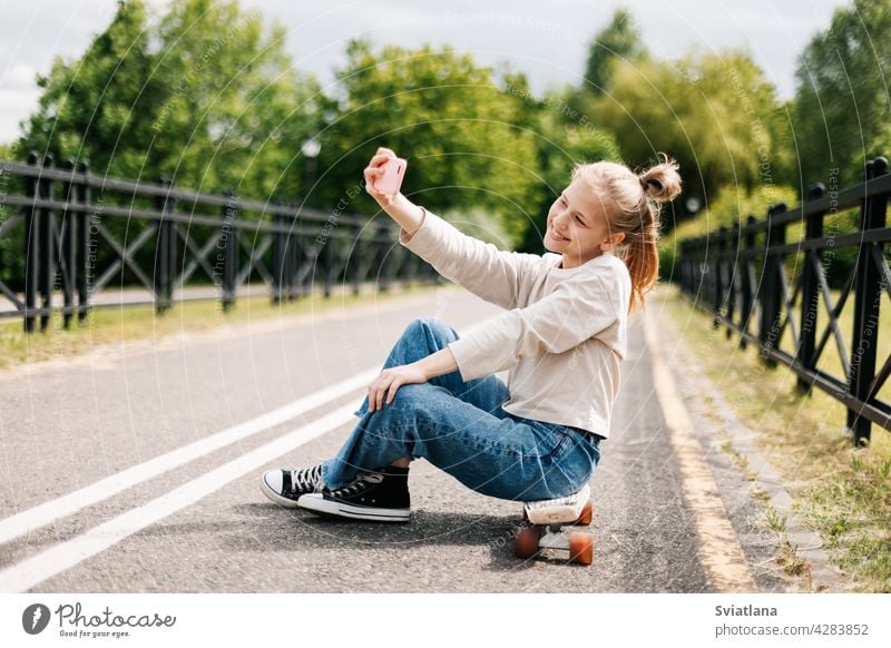 Nettes blondes Teenager-Mädchen, das auf einem Skateboard in einem Stadtpark sitzt, mit Freunden auf einem Smartphone chattet und ein Selfie macht.