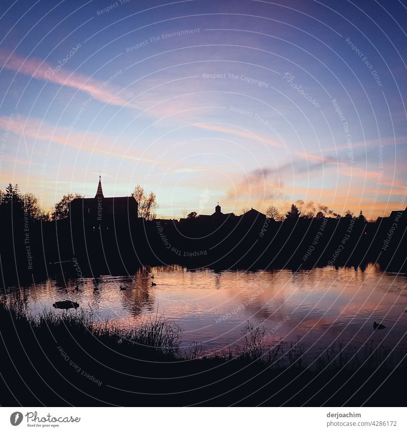 Kleine Fränkische Stadt im Abendlicht. Strukturen der Kirche sowie der Häuser sind zu sehen. Im Vordergrund ein Karpfenweiher in dem sich der noch blaue Himmel und weiße Streifen abzeichnen. .