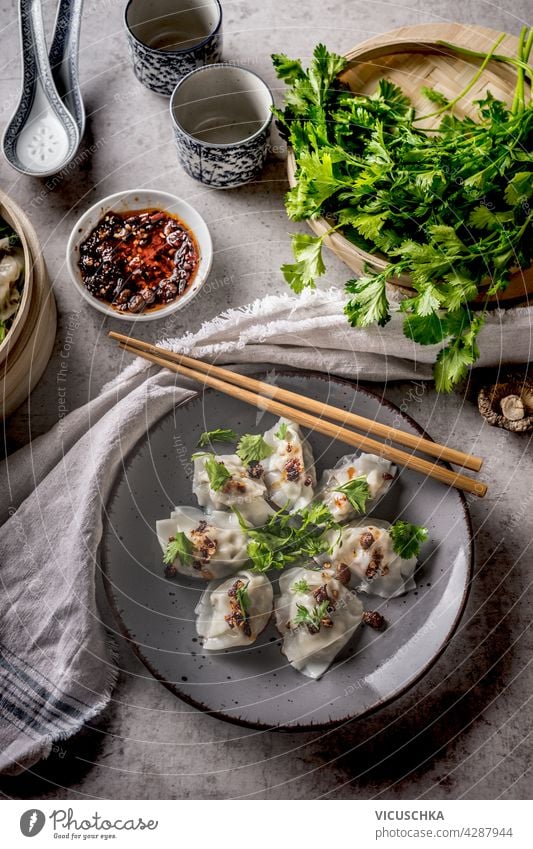 Asiatische Knödel serviert mit traditionellem Geschirr, Stäbchen, Bündel Koriander und Chilisauce. Grauer Beton-Hintergrund. Ansicht von oben Textfreiraum