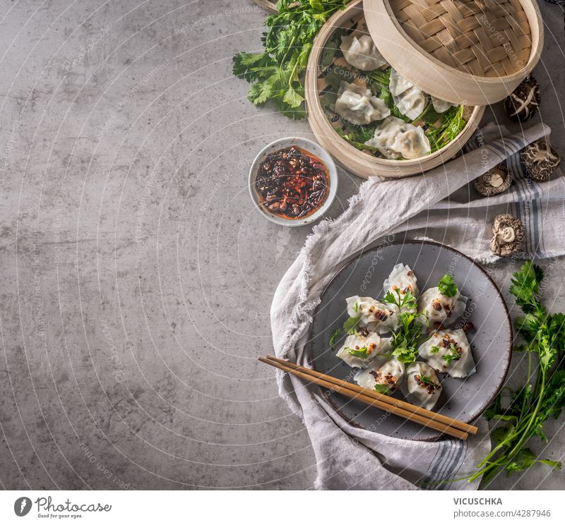 Asiatisches Essen Konzept mit hausgemachten Knödeln auf Teller und typische Ausrüstung und Zutaten wie Stäbchen, Dampfer, frische asiatische Kräuter und Chili-Sauce. Dunkler Beton-Hintergrund. Ansicht von oben. Essen Grenze