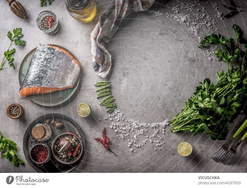 Frischer roher Lachs mit Rahmen von Gewürzen, Öl, frischen Kräutern und Zitrone auf grauem Beton Hintergrund. Kochen Vorbereitung mit frischen Zutaten zu Hause. Ausrüstung: Gabel, Schüsseln und Küchentuch. Ansicht von oben