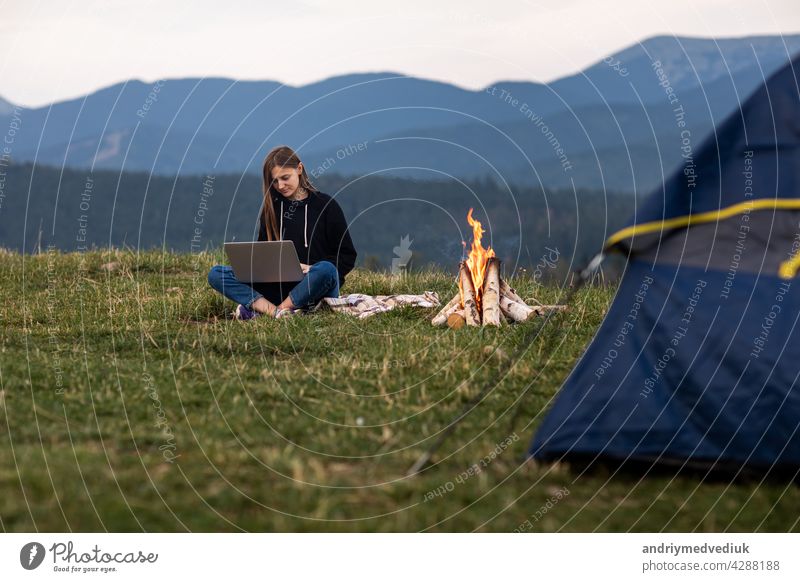 junge weibliche Freiberufler arbeiten am Laptop in den Bergen am Abend. Tourist Mädchen sitzt in der Nähe von Lagerfeuer und Zelt. Kopierraum Sitzen Frau Urlaub