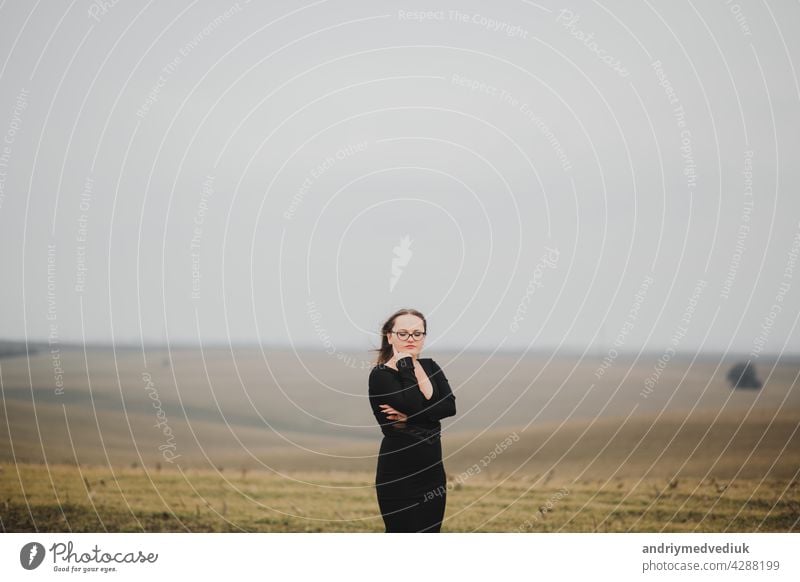 Junges Mädchen 20-25 Jahre alt mit Brille und einem schwarzen Kleid posiert auf einem Feld Hintergrund Frau jung im Freien Schönheit modisch Pose Natur Model